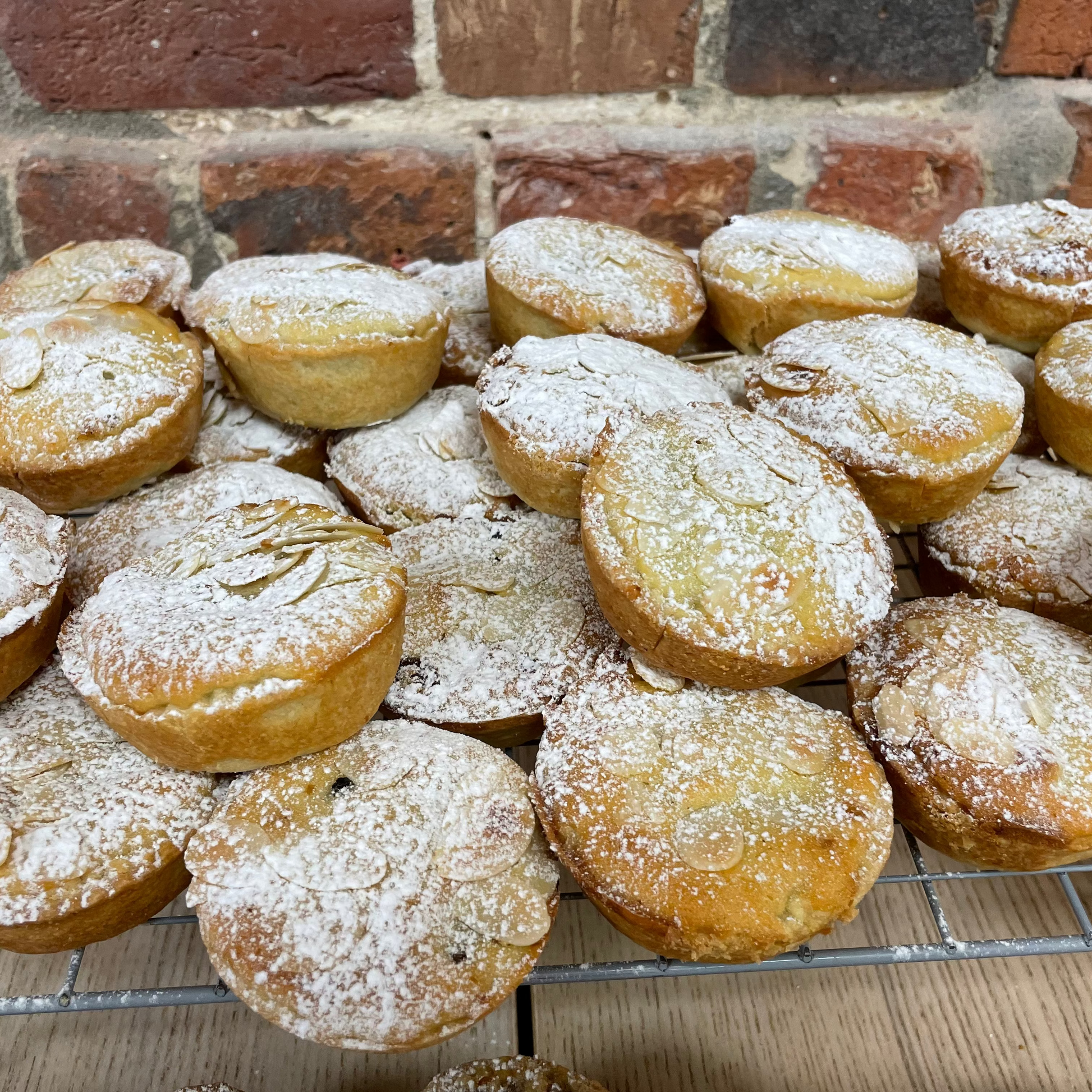 Piles of frangipane mince pies dusted with icing sugar on a metal rack against a brick wall.